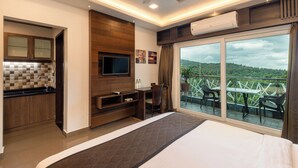 A modern hotel room with a bed, a TV, a balcony with outdoor seating, and a kitchen area.