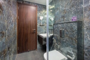 Une salle de bain avec un mur en marbre, une porte en bois, une cabine de douche en verre, des toilettes blanches et un lavabo avec un miroir.