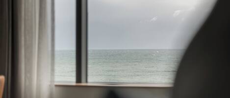 A view through a window with a curtain, revealing a body of water and a cloudy sky.