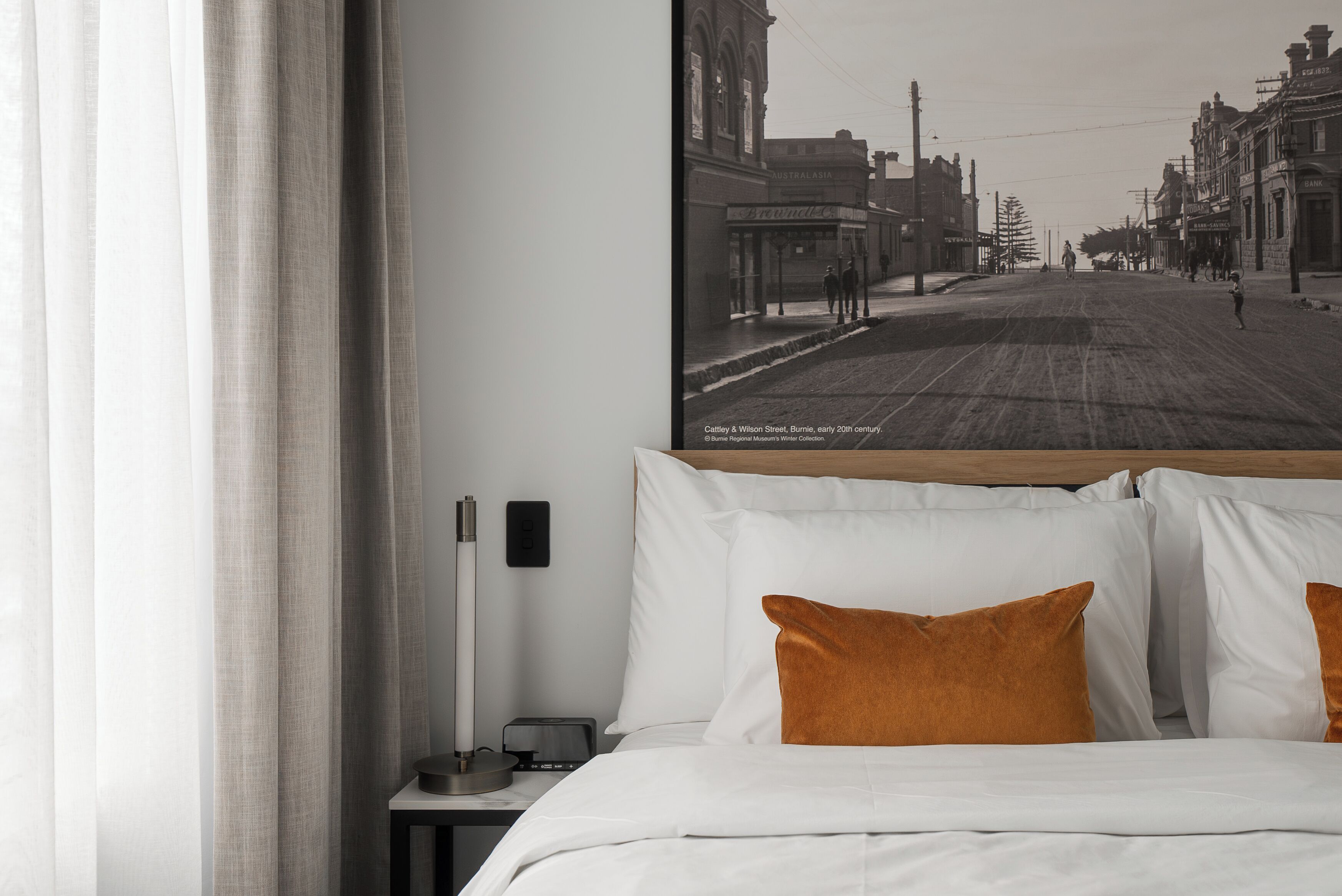 Ein Hotelzimmer mit einem großen historischen Foto an der Wand, einem Bett mit weißen und orangefarbenen Kissen und einem Nachttisch mit Lampe.