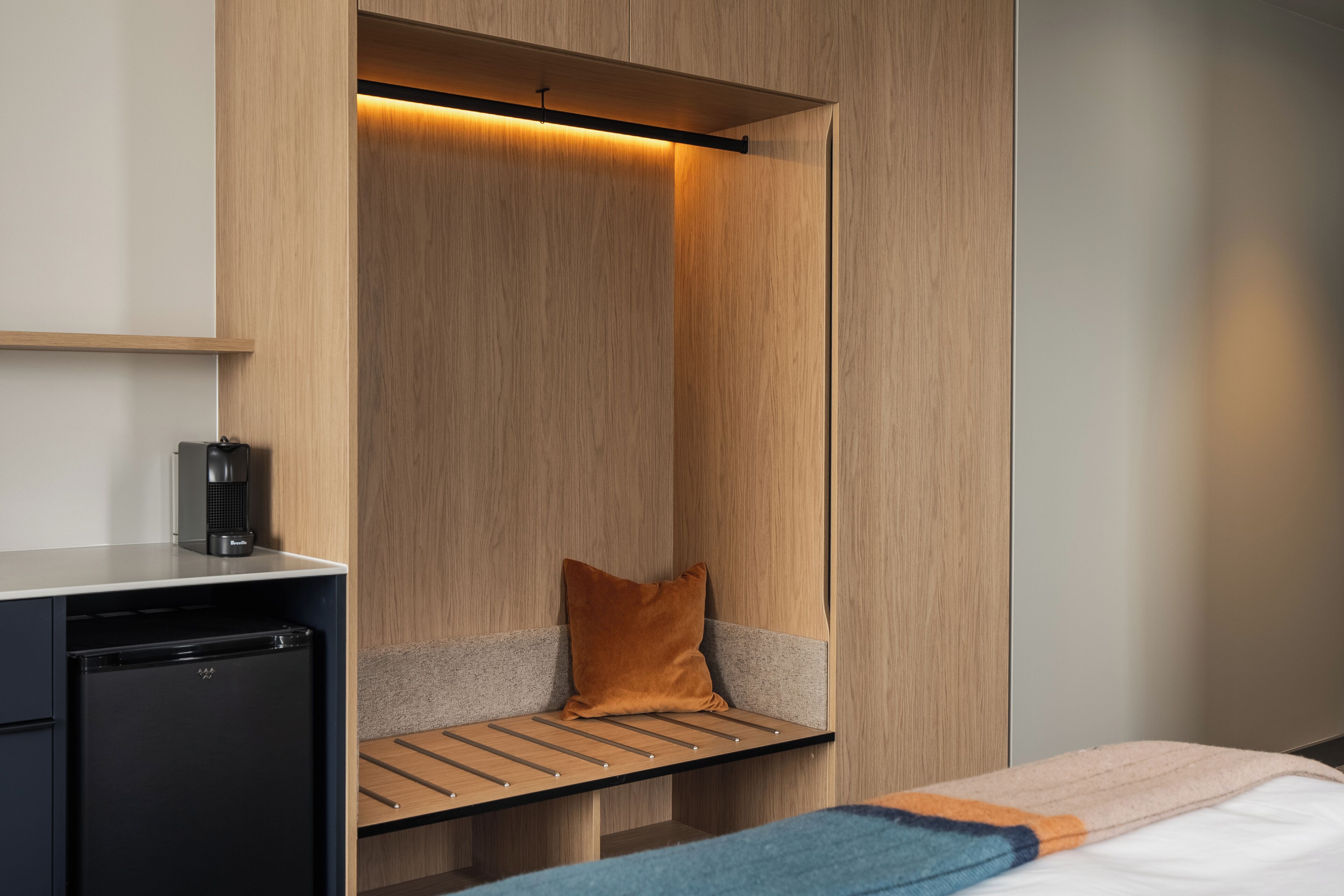 A compact hotel room with a built-in desk, a small seating area with a bench and a pillow, and a bed with a blanket.