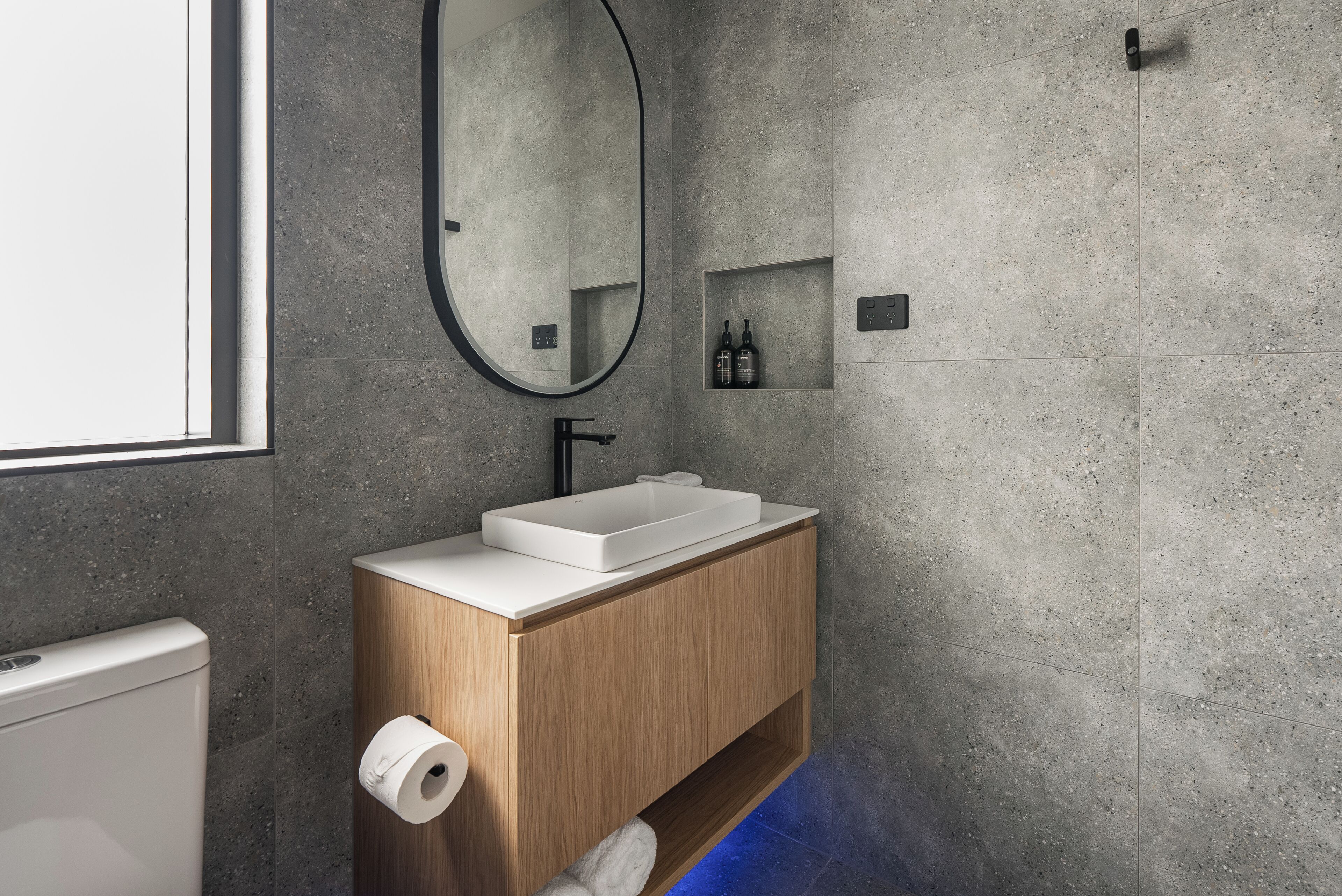 Un baño moderno con un lavamanos rectangular, tocador de madera, un espejo y un estante pequeño con artículos de aseo.