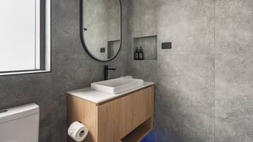 Un baño moderno con un lavamanos rectangular, tocador de madera, un espejo y un estante pequeño con artículos de aseo.