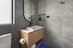 Un baño moderno con un lavamanos rectangular, tocador de madera, un espejo y un estante pequeño con artículos de aseo.