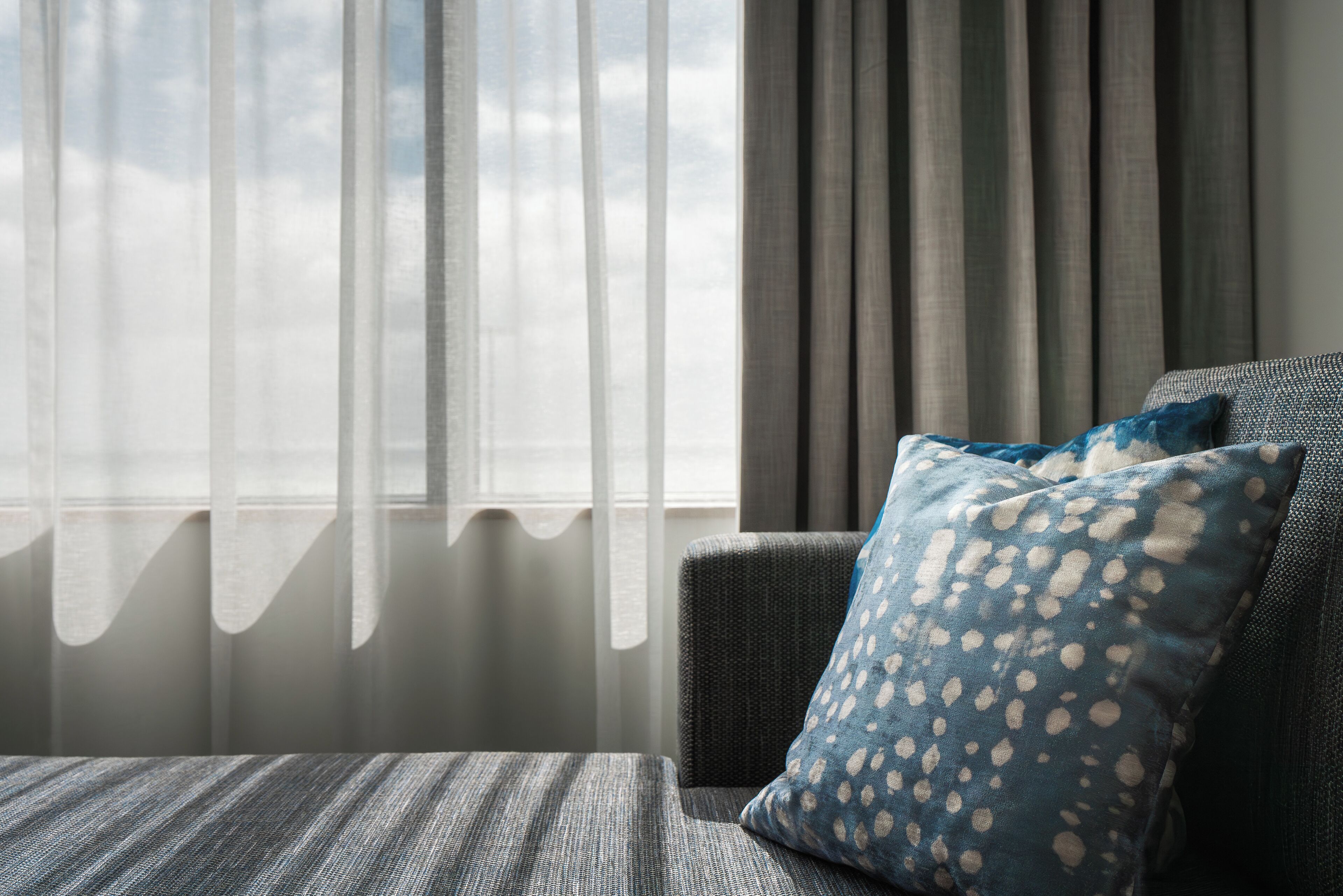 Una habitación con vistas, cortinas translúcidas, un sofá gris con un cojín estampado y una ventana que da al cielo.