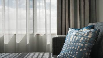 A room with a view through sheer curtains, a grey sofa with a patterned pillow, and a window with a view of the sky.