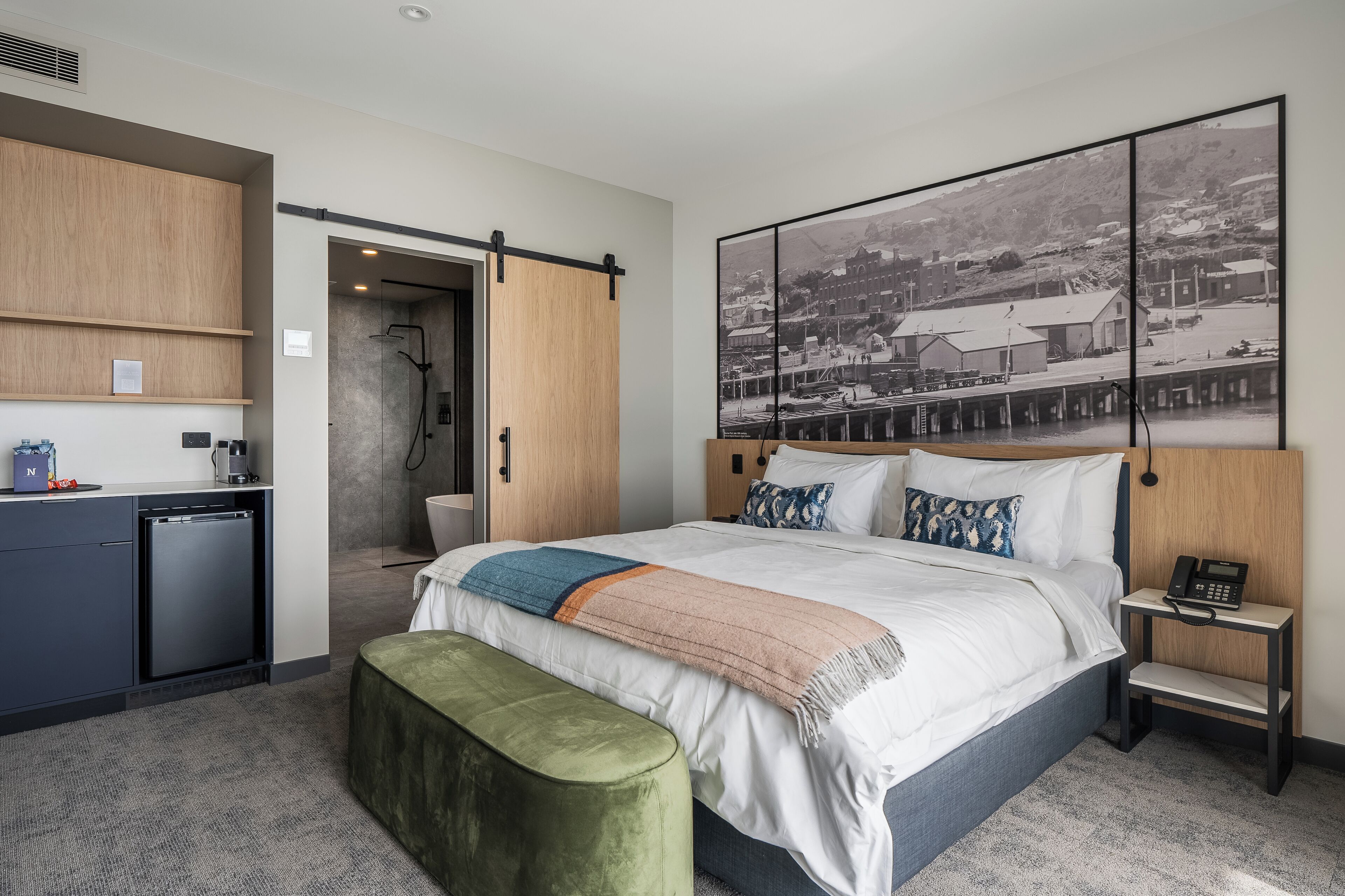 Habitación de hotel moderna con cama, un escritorio pequeño, ducha y un ventanal grande.