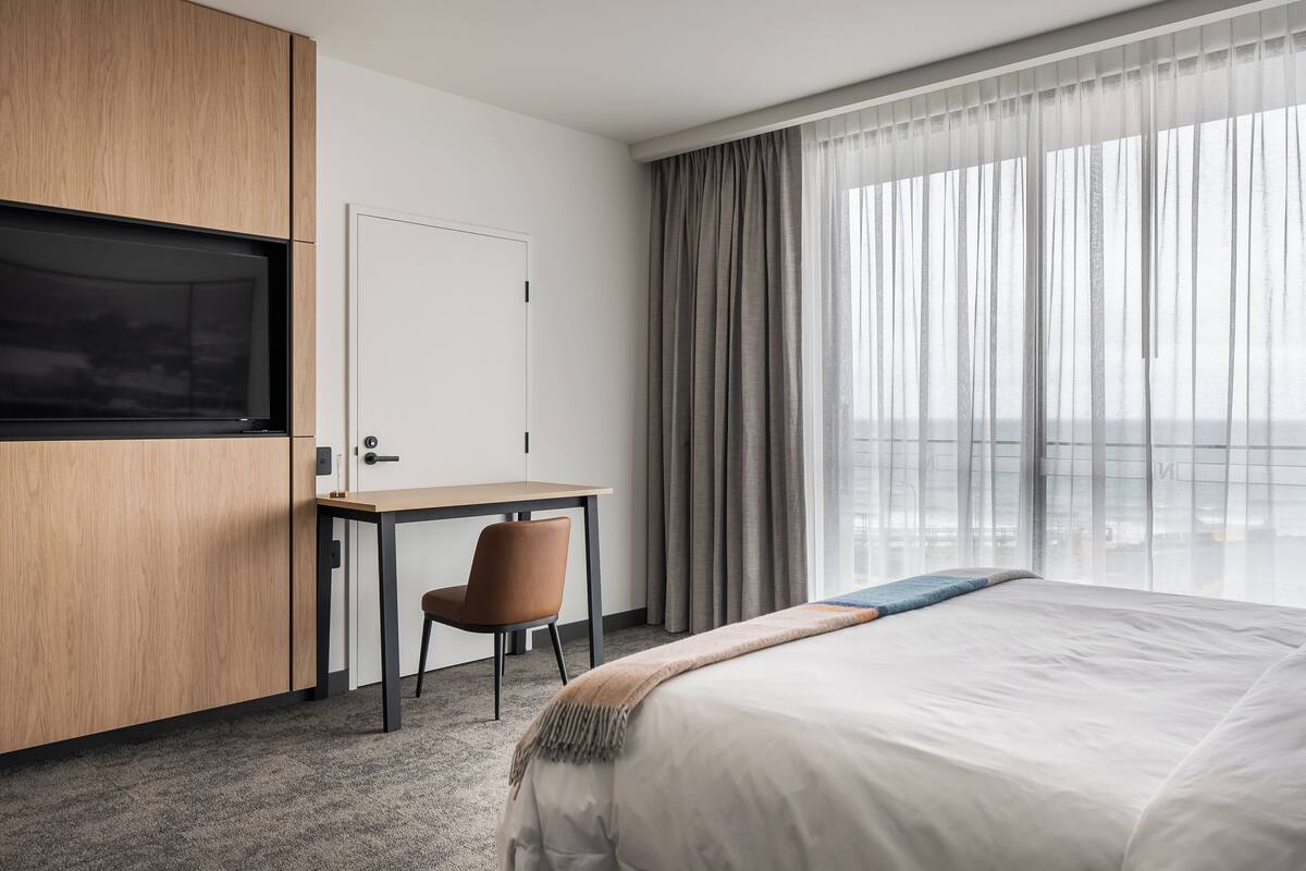Quarto de hotel com cama, escrivaninha, cadeira, televisão e uma grande janela com cortinas.