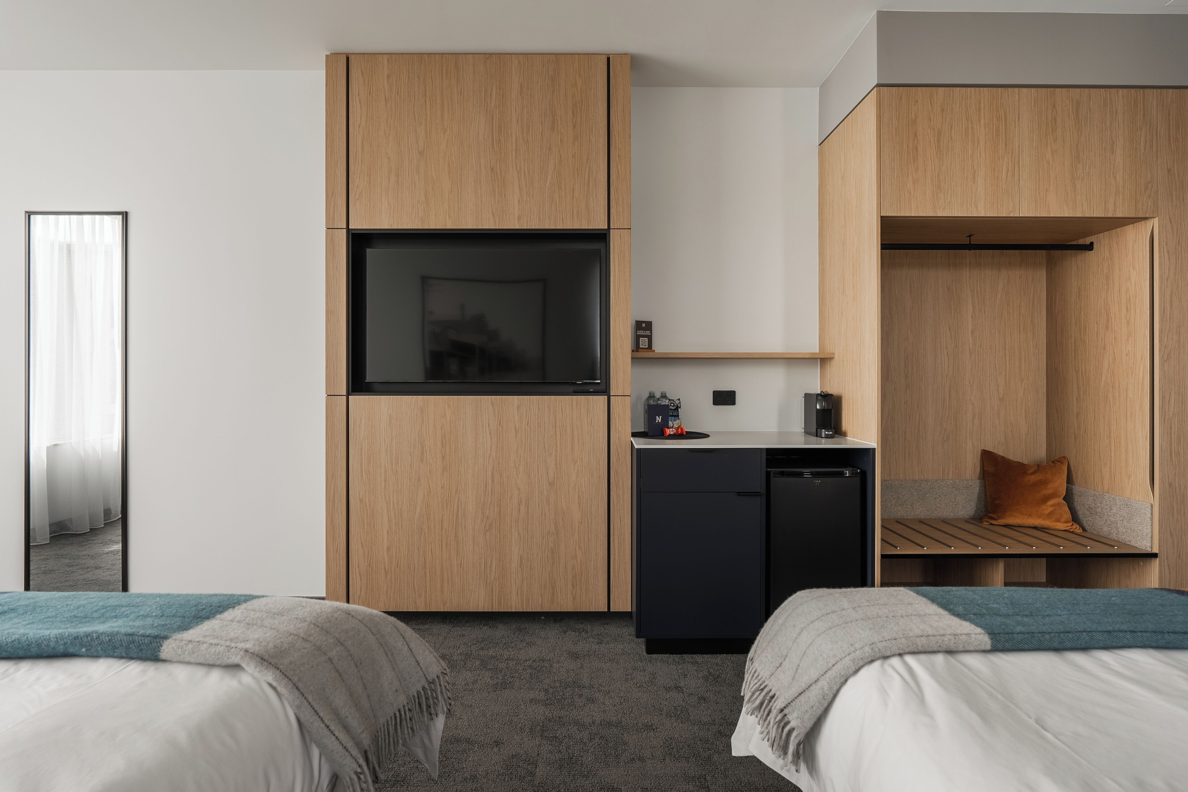 A modern hotel room with two beds, a built-in desk, a small kitchenette, and a large mirror.