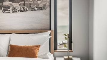 A hotel room with a bed, a nightstand, a lamp, and a large window with a view of a beach and vehicles.