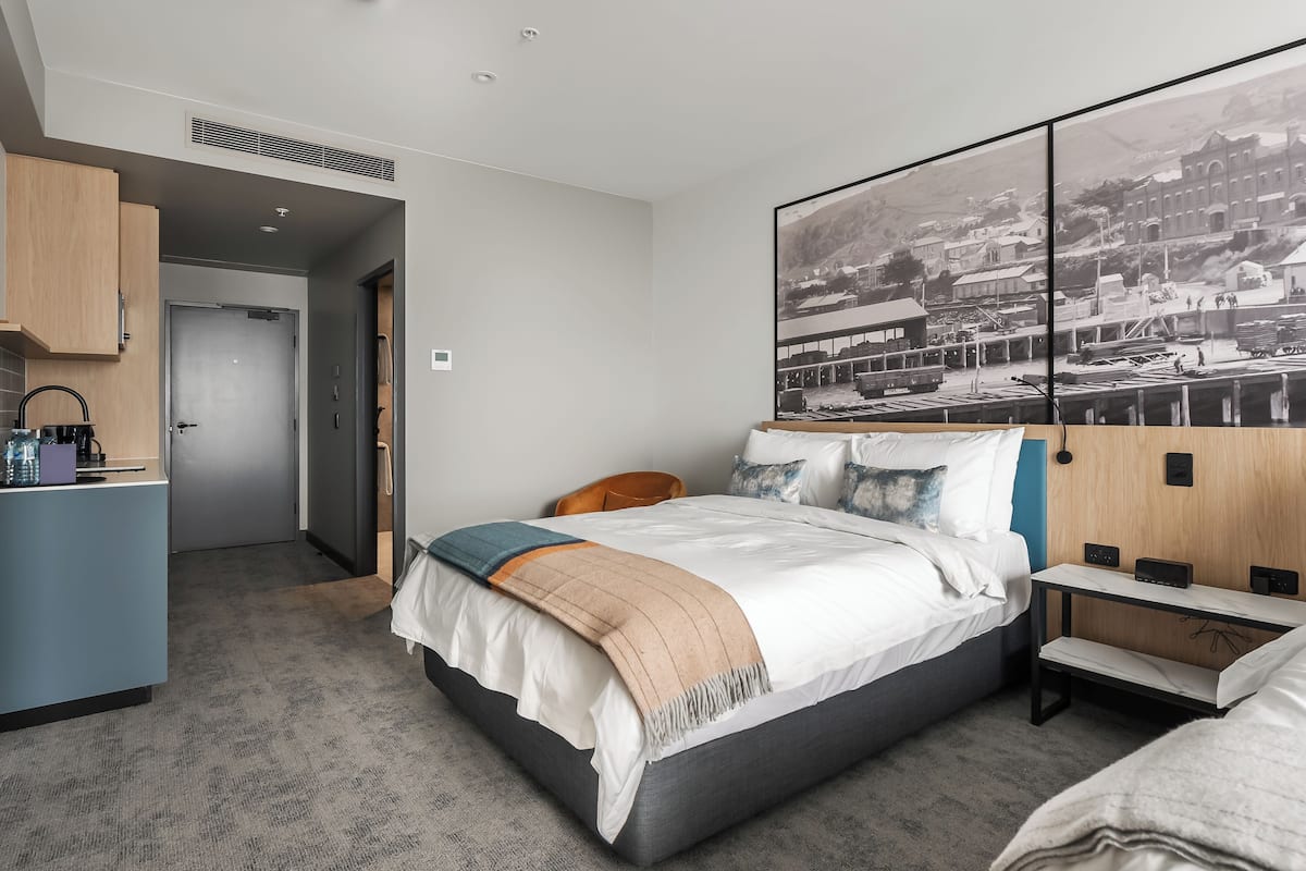 Quarto de hotel moderno com uma cama, mesa de cabeceira e uma grande pintura em preto e branco retratando uma paisagem urbana.