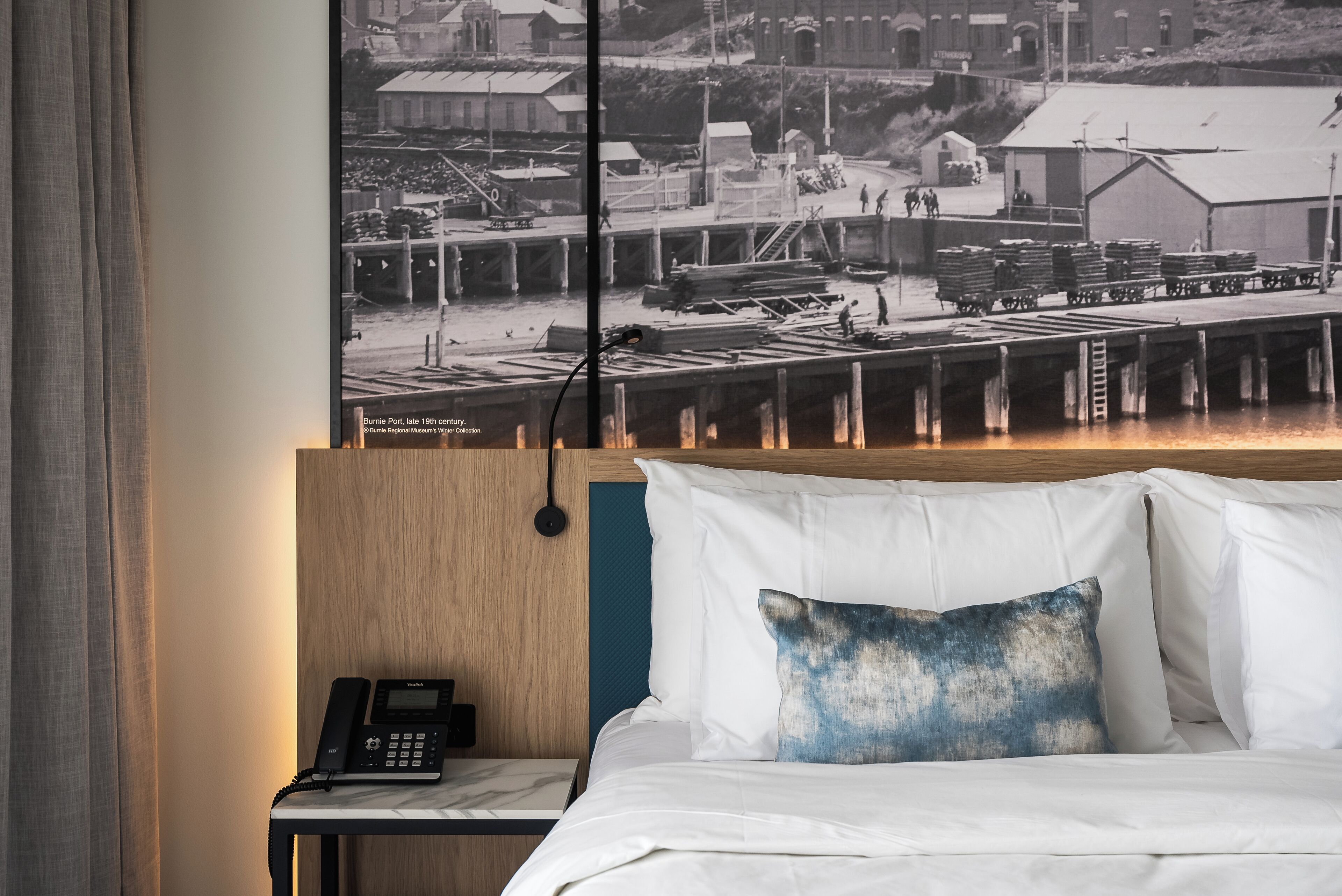 A hotel room with a bed, a nightstand with a telephone, and a wall-mounted picture of a historical dock scene.