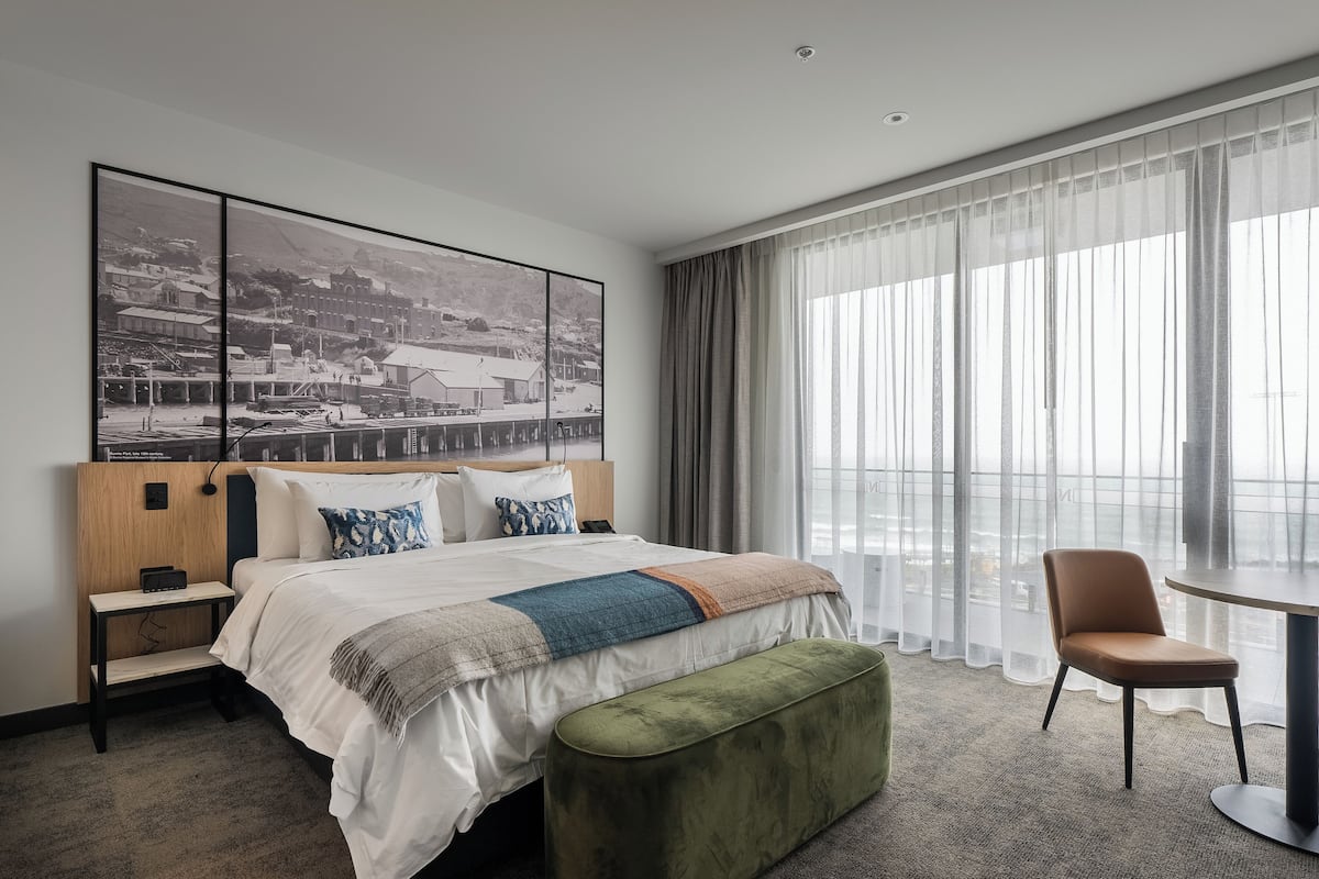 Quarto de hotel com uma cama grande, uma mesa de cabeceira, um banquinho verde, uma cadeira e uma janela ampla com cortinas leves.