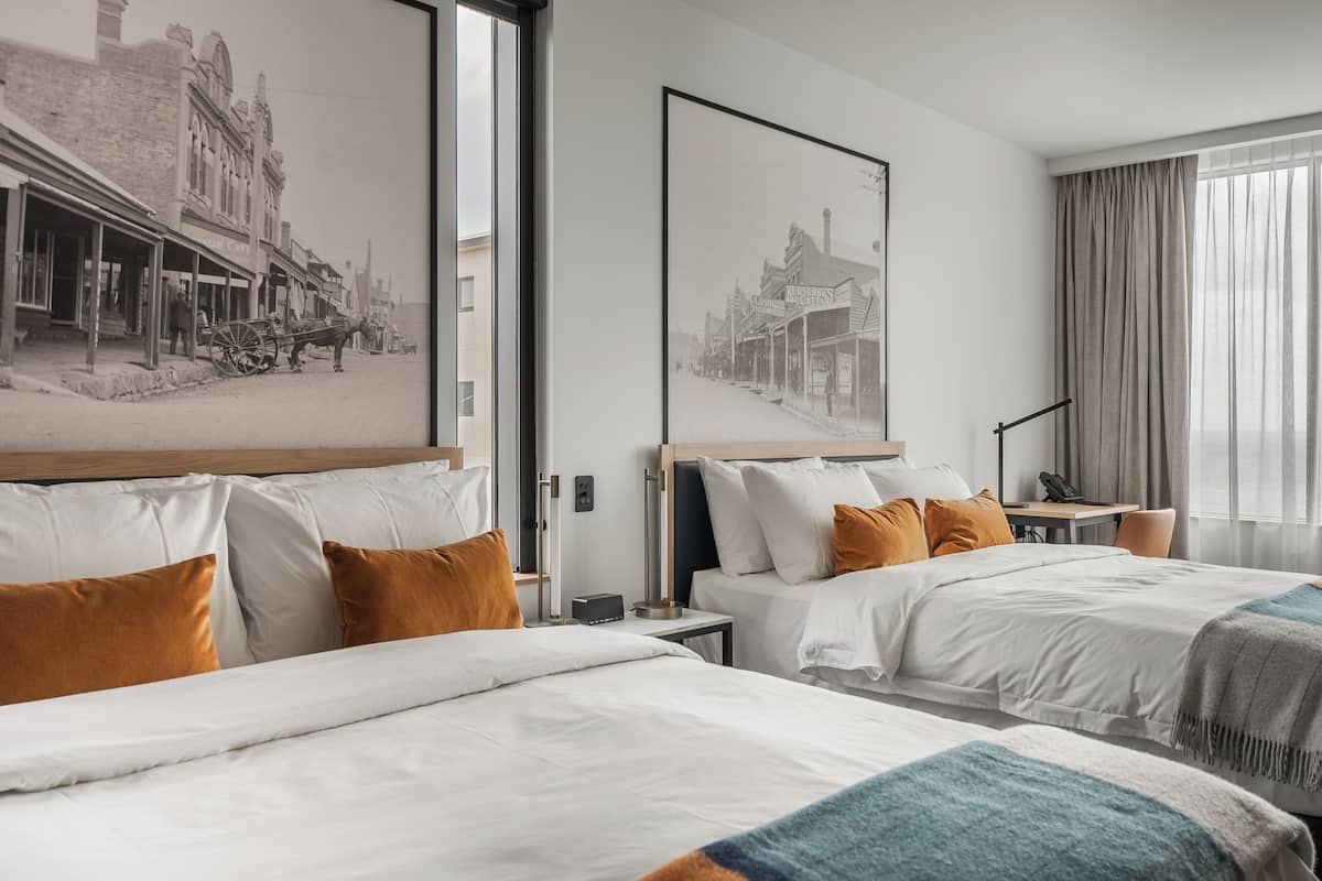 Quarto de hotel com duas camas, uma janela grande e uma representação histórica de uma rua emoldurada na parede.