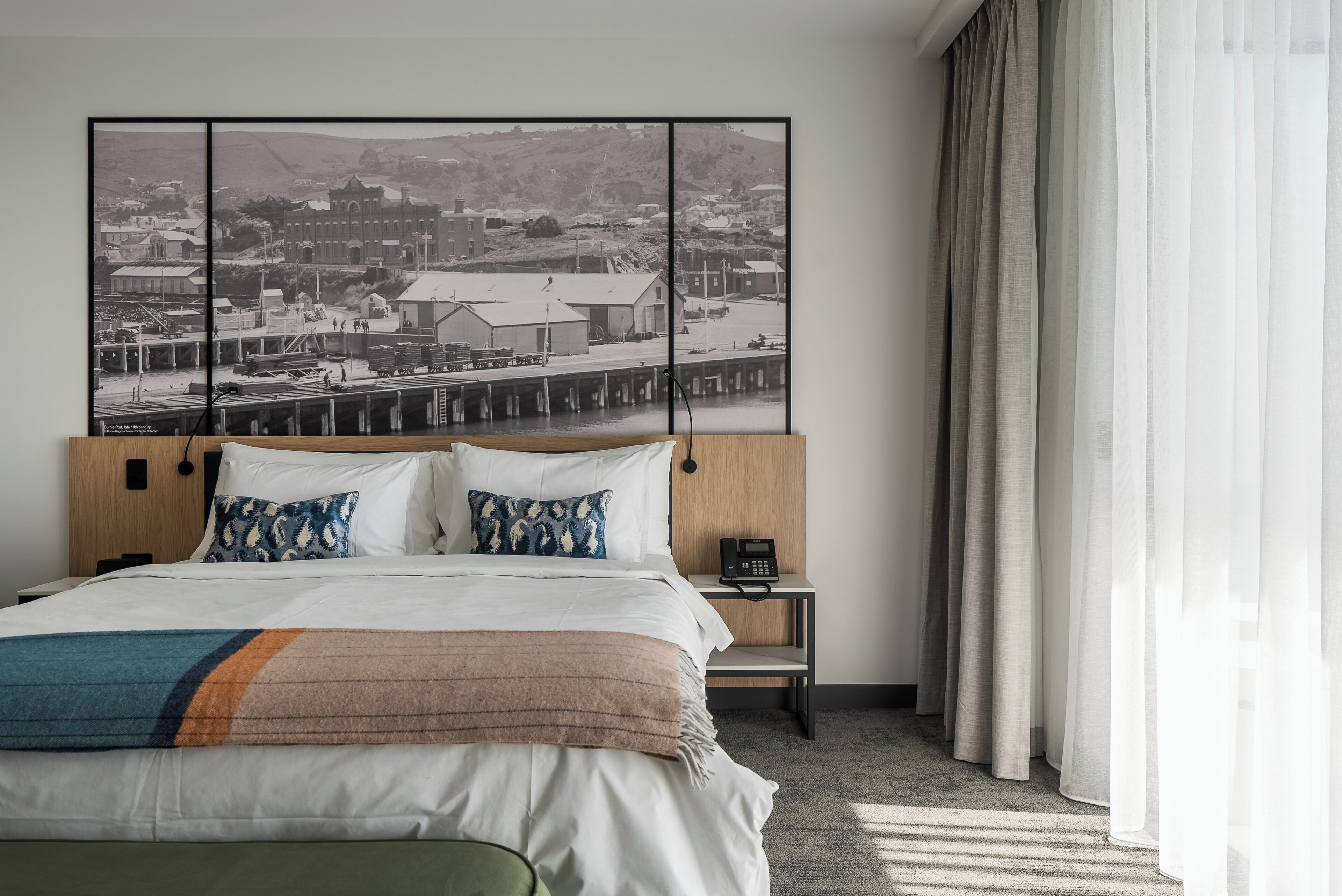Un cuarto de hotel con una cama grande, un cabecero de madera, una mesita de noche con un teléfono y una fotografía grande en blanco y negro en la pared.