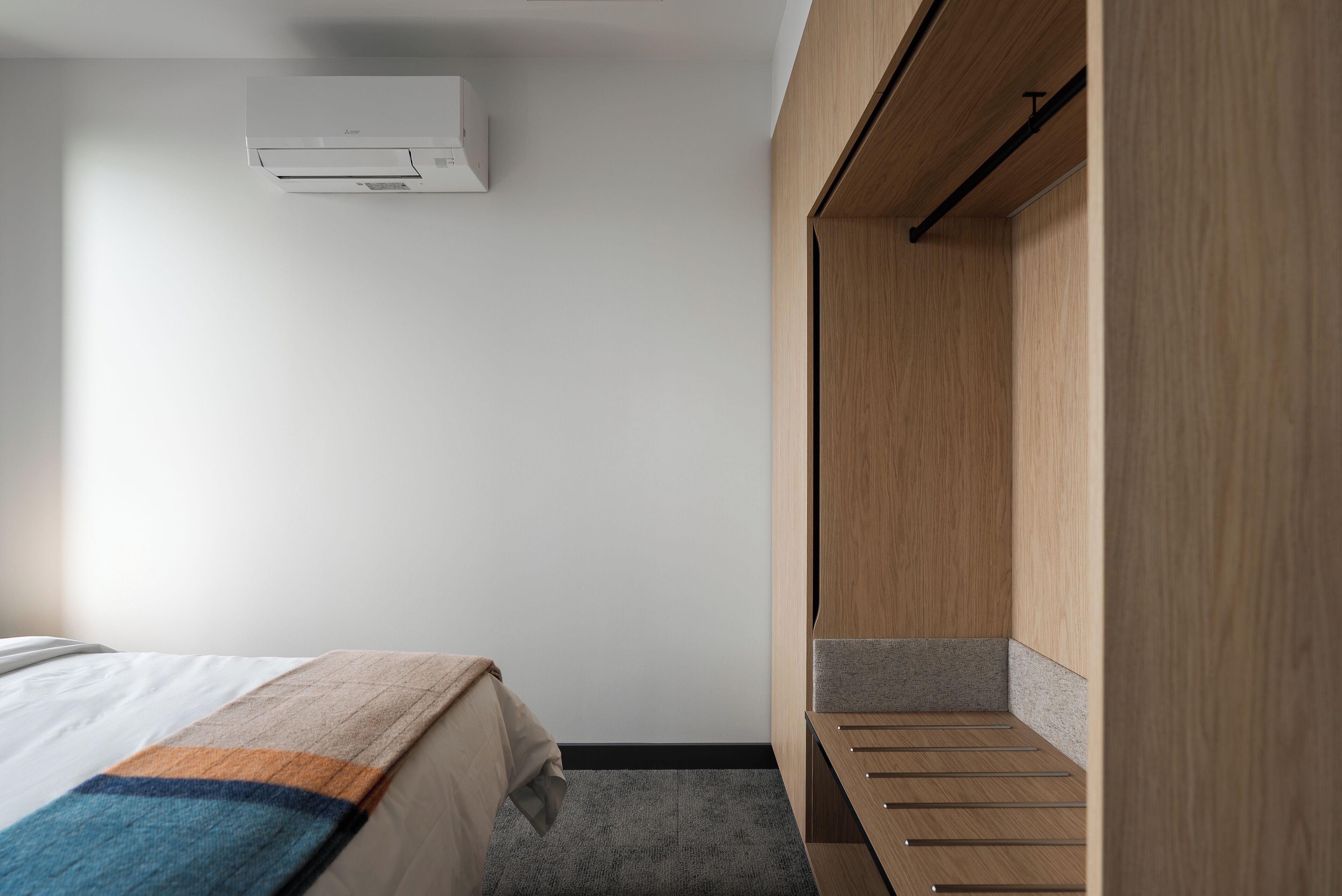 Una habitación de hotel con una cama, un armario de madera, una alfombra gris y un aire acondicionado blanco en la pared.