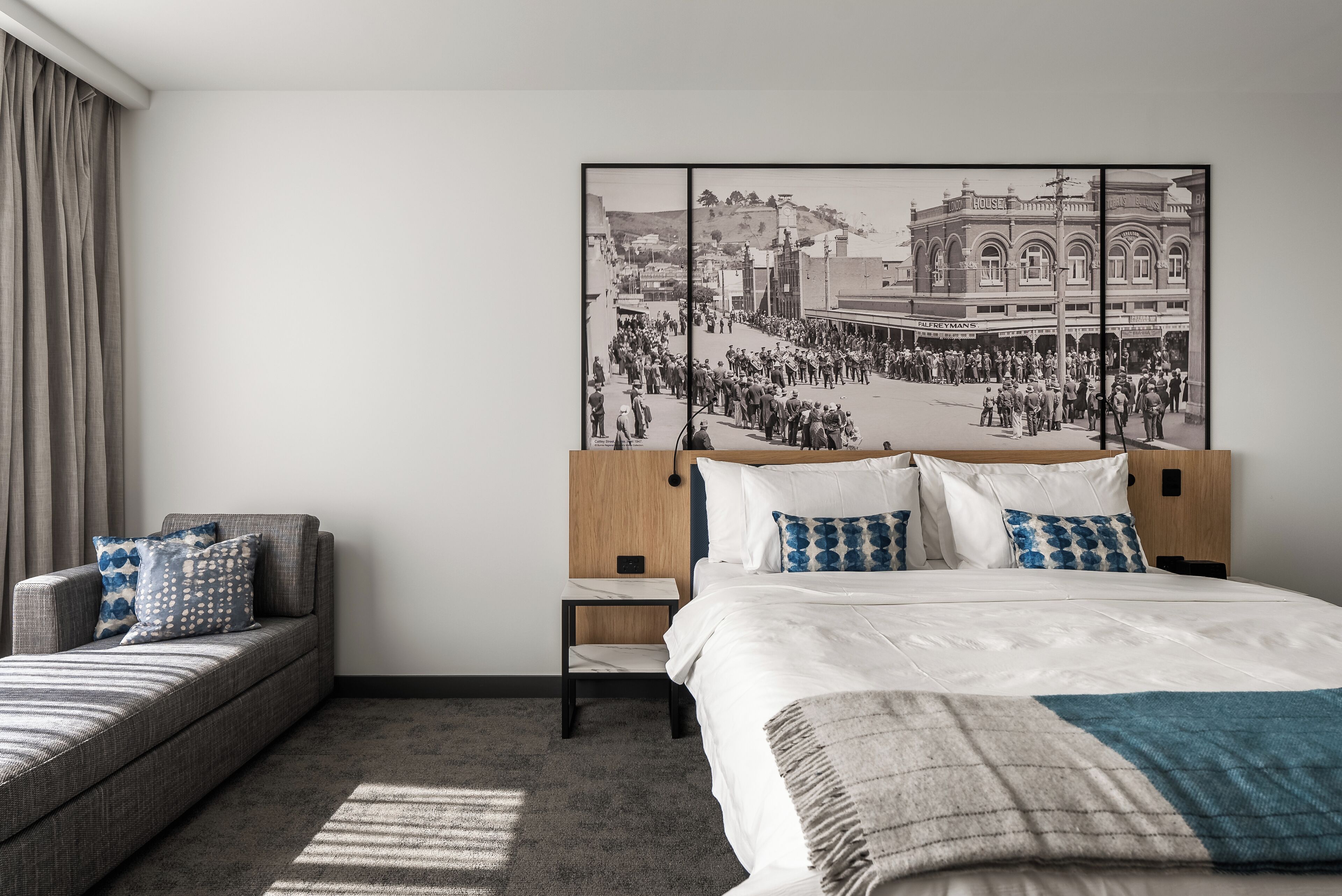 Una habitación de hotel moderna con una cama grande, un sofá, una mesita de noche y un cuadro colgado en la pared que muestra una escena callejera histórica.