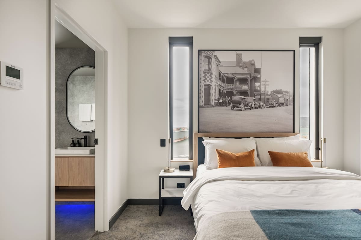 Quarto de hotel com uma cama, mesa de cabeceira, espelho, fotografia emoldurada na parede e vista para a rua.