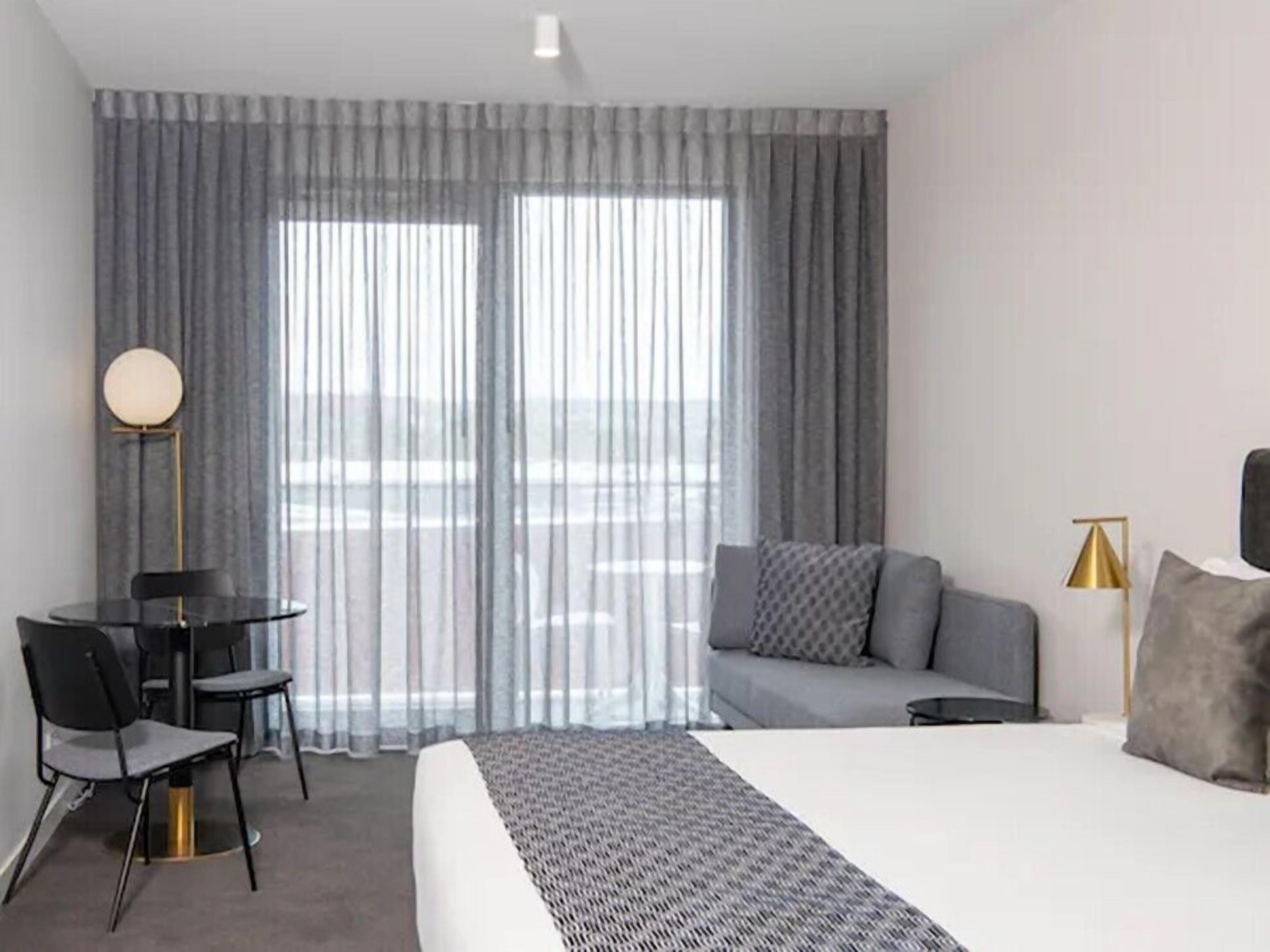 A hotel room with a bed, a sofa, a desk with chairs, a lamp, and large windows with curtains.