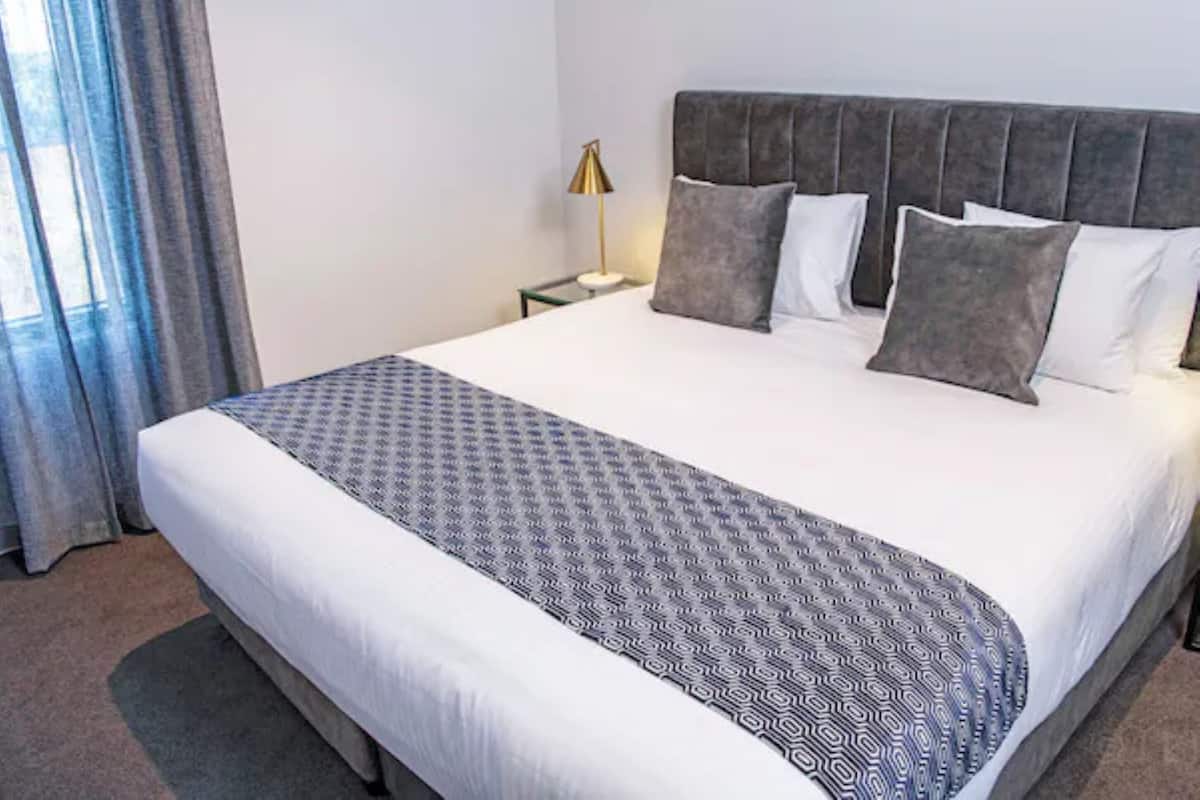 Quarto de hotel com uma cama grande, mesa de cabeceira, abajur e vista para o exterior através de uma janela com cortinas.