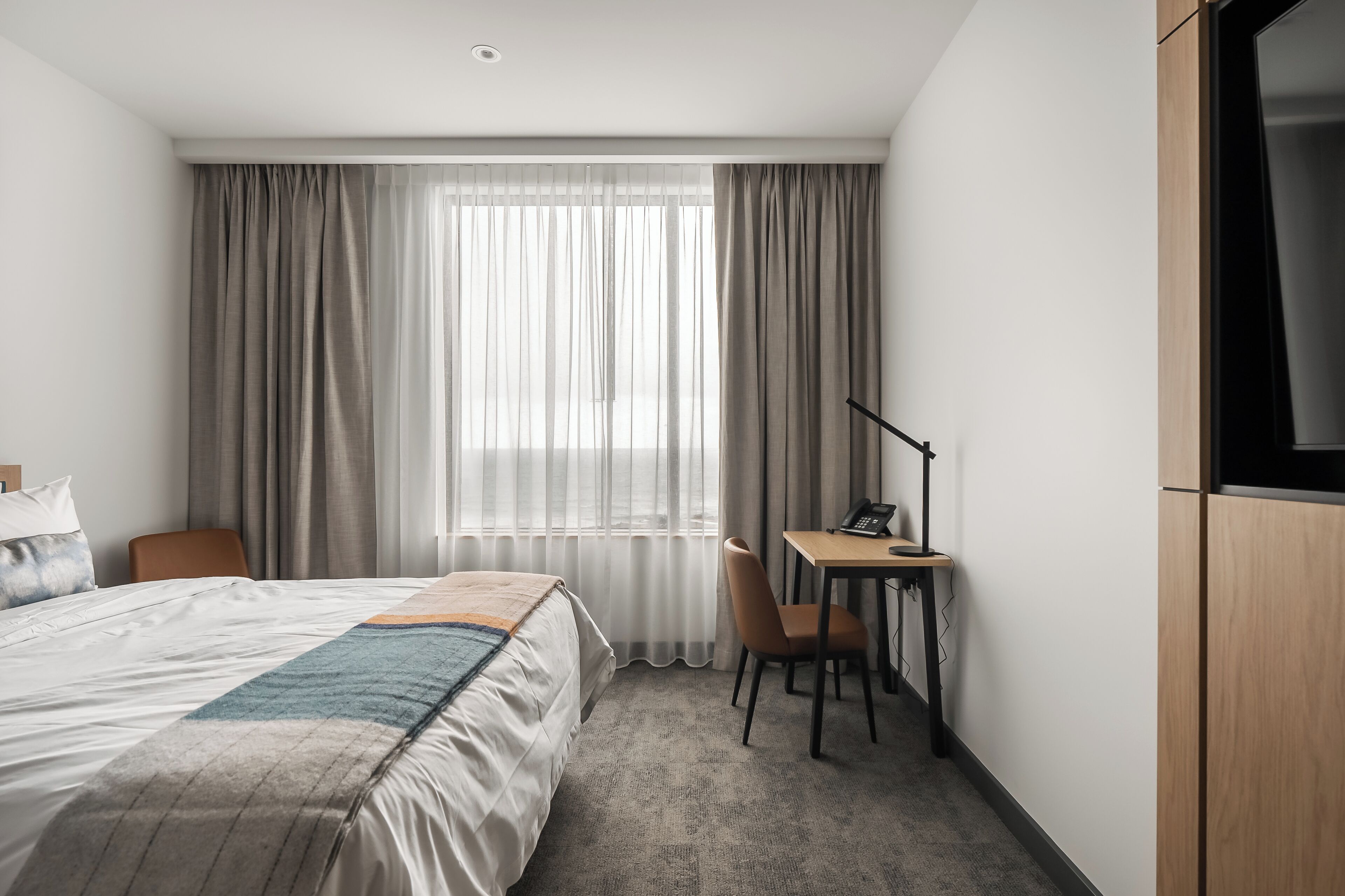 Et hotelværelse med seng, skrivebord, stol og fjernsyn.