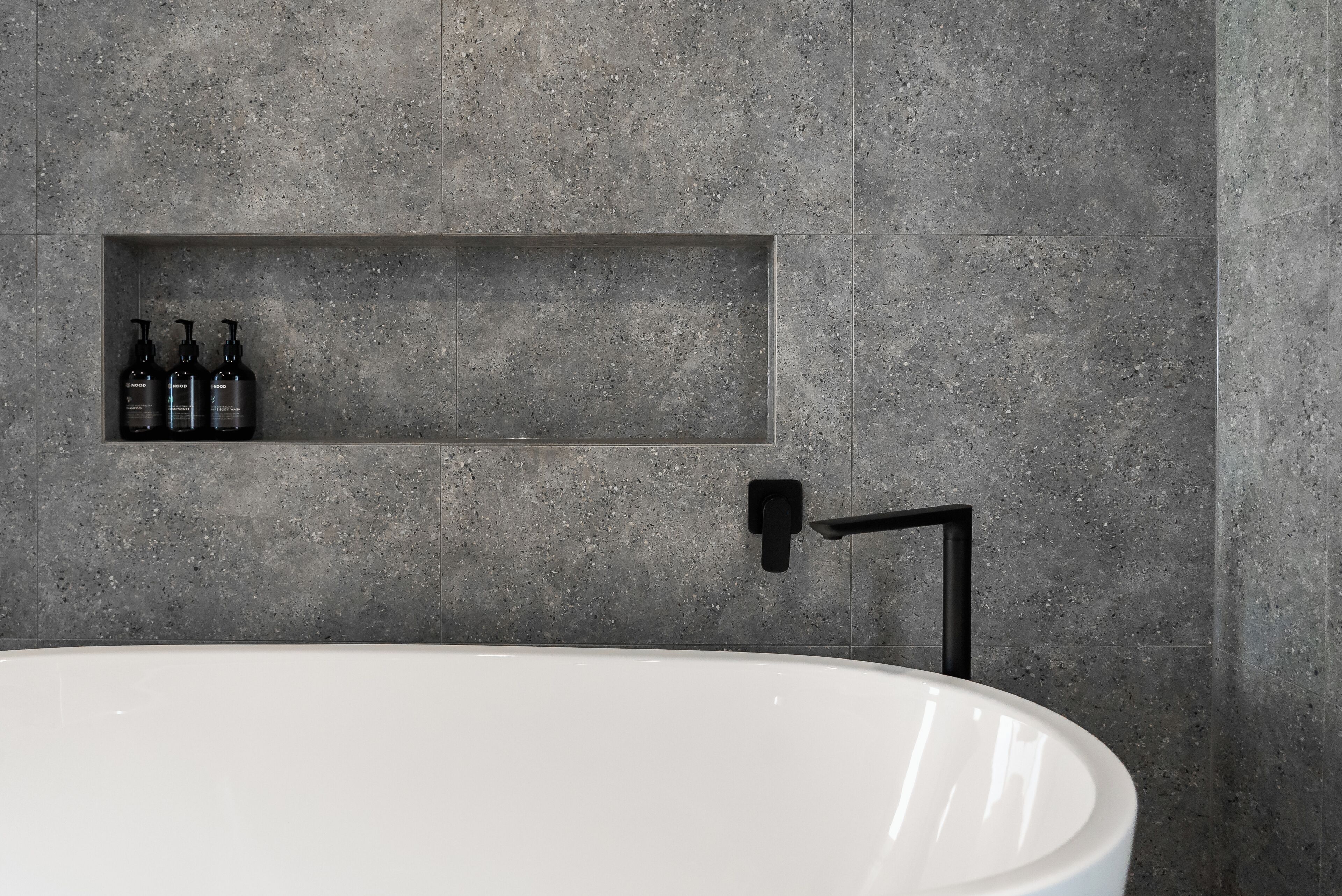 A modern bathroom with a freestanding bathtub, a built-in shelf with toiletries, and a black faucet.