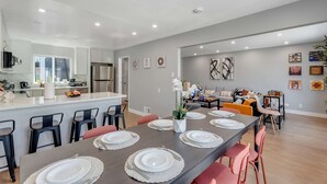 A modern kitchen with a central island, dining area with a long table set for six, and a living room with a sofa and coffee table.