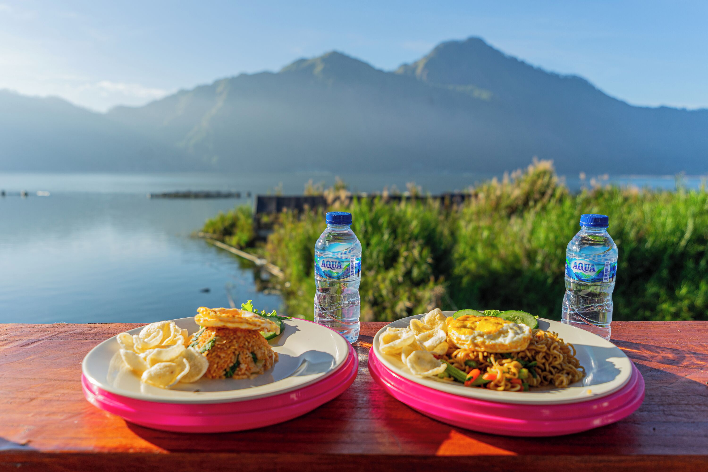 Two plates of food on a wooden table with a view of a lake and mountains in the background.