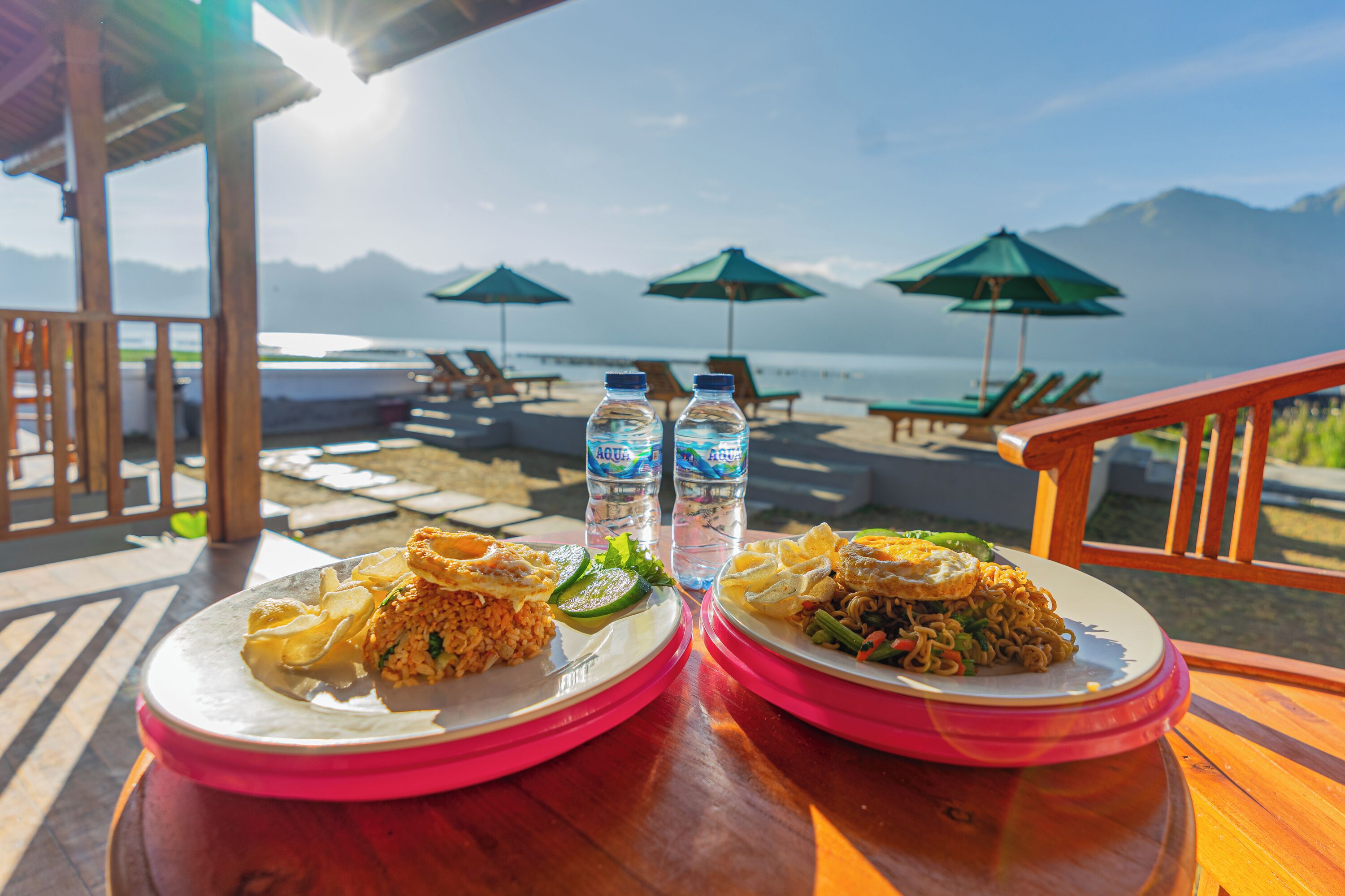 Two plates of food on a wooden table with a view of a beach and mountains in the background.