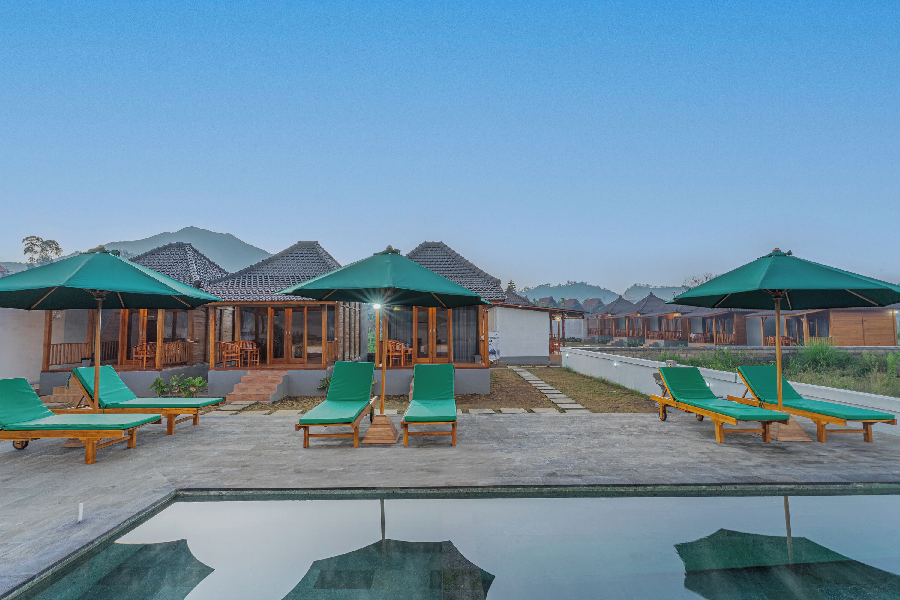 A poolside area with lounge chairs and umbrellas, wooden buildings with large windows, and a clear sky.