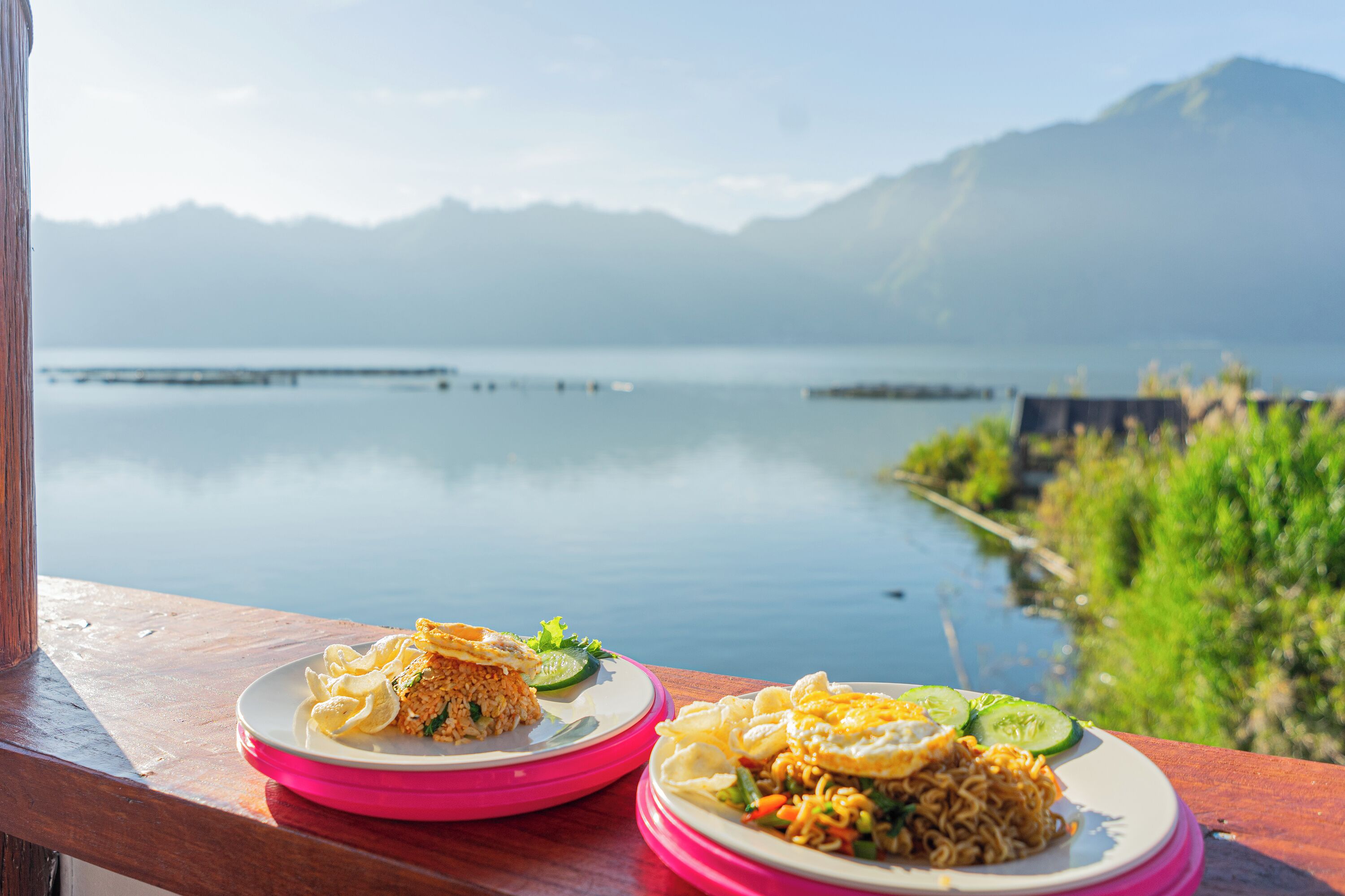 Two plates of food on a wooden table with a view of a calm lake and mountains in the background.