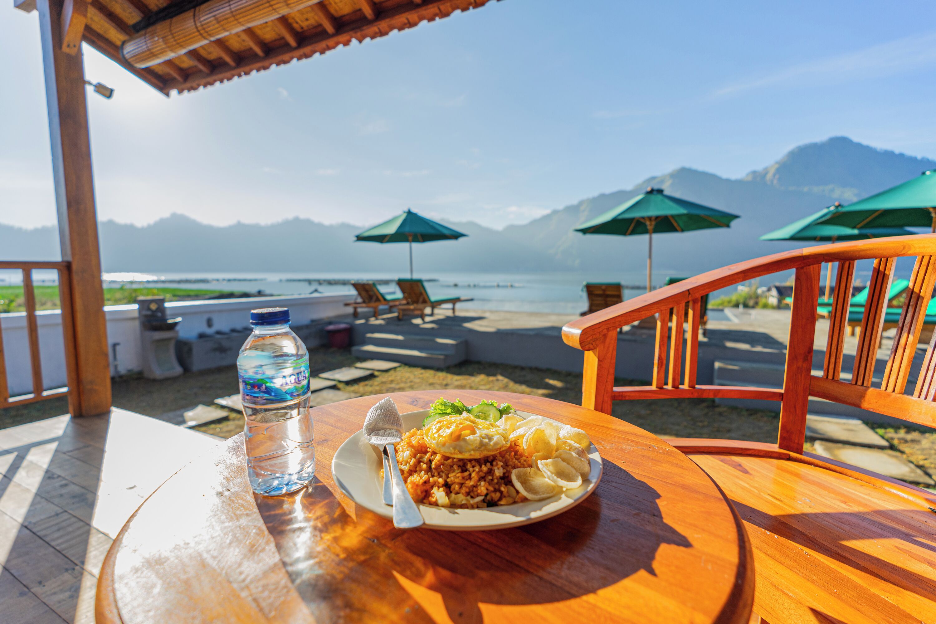 Een houten tafel met een bord eten en een waterfles, met uitzicht op een strand met parasols en stoelen.