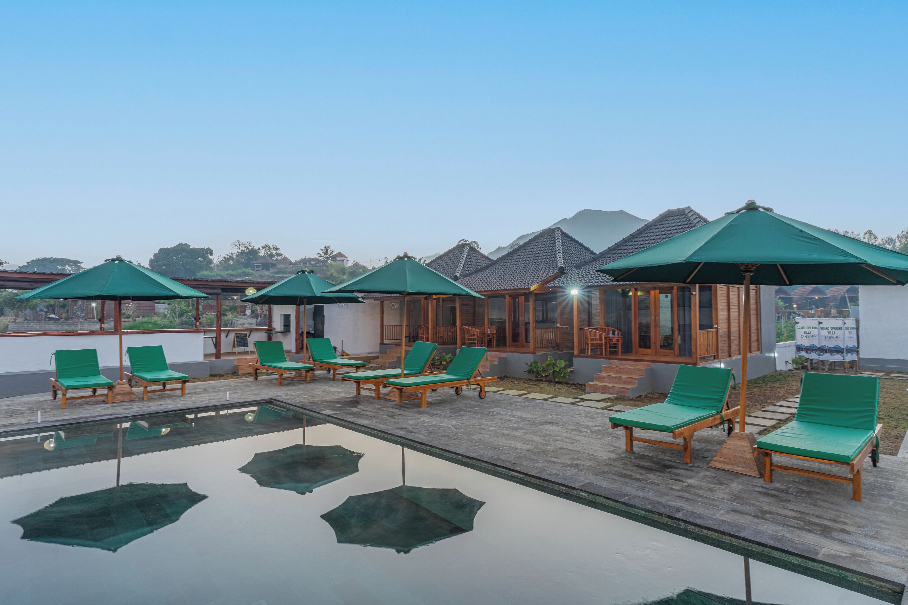A rooftop pool area with green lounge chairs and umbrellas, wooden deck, and a view of mountains in the background.
