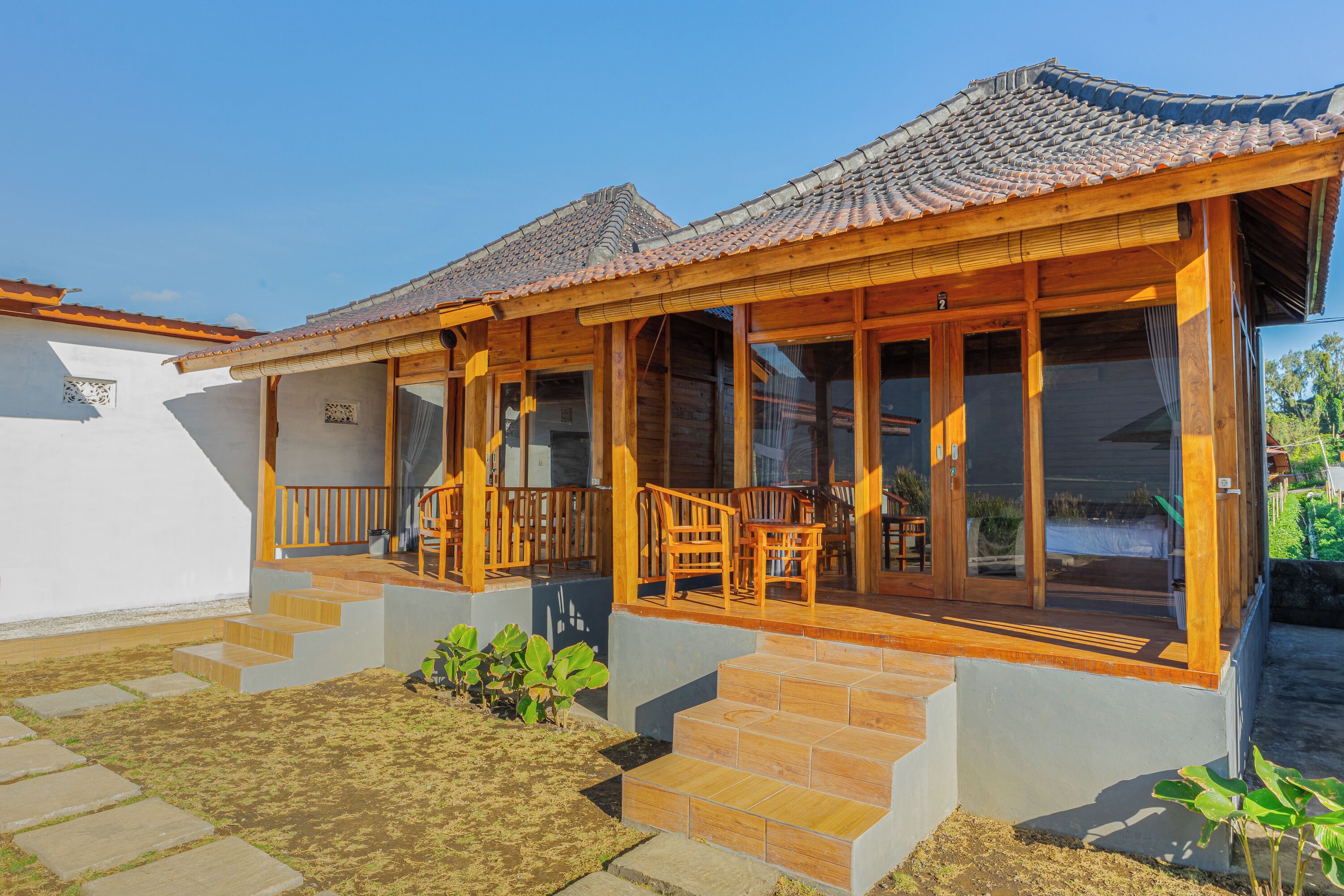 A traditional wooden house with a tiled roof, a porch with wooden chairs, and a staircase leading to the entrance.