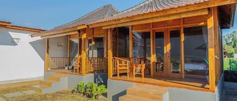 A traditional wooden house with a tiled roof, a porch with wooden chairs, and a staircase leading to the entrance.