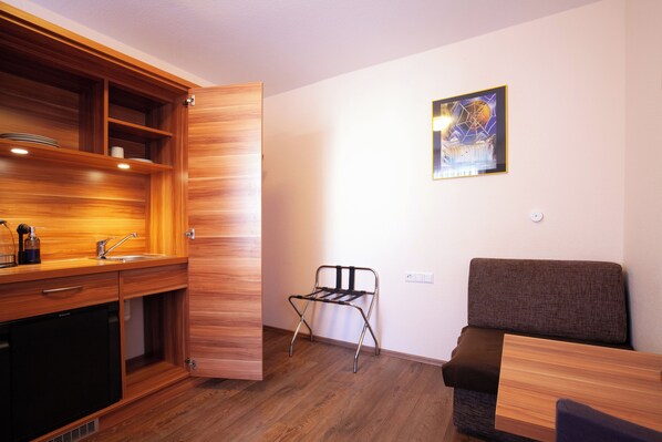 A compact kitchen with wooden cabinets, a sink, and a compact refrigerator. There is a foldable chair and a sofa in the living area. - Hotel Garni Zum Wilden Mann (Künzelsau)
