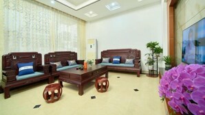 A spacious living room with a sofa set, a coffee table, and a small stool. There are potted plants and a large window with patterned curtains. - Mount Danxia Xin Yue Inn (Renhua)