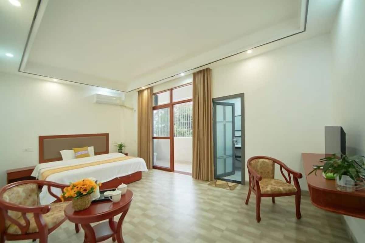 A hotel room with a bed, a desk, a chair, a small table with a vase of flowers, and a sliding glass door leading to an outdoor area.