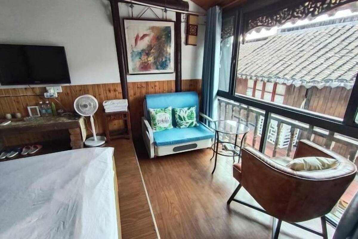 A room with a bed, a blue sofa with cushions, a brown armchair, a small table, a fan, and a TV.