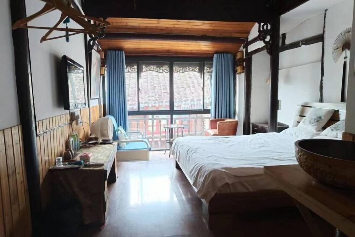 A hotel room with a large bed, a wooden headboard, a table with a chair, a window with curtains, and a balcony with a view of buildings.