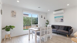 A bright living room with a grey sofa, a dining table with chairs, a potted plant, and a large window offering a view of greenery. - Life's a Beach Shack (Cowes)