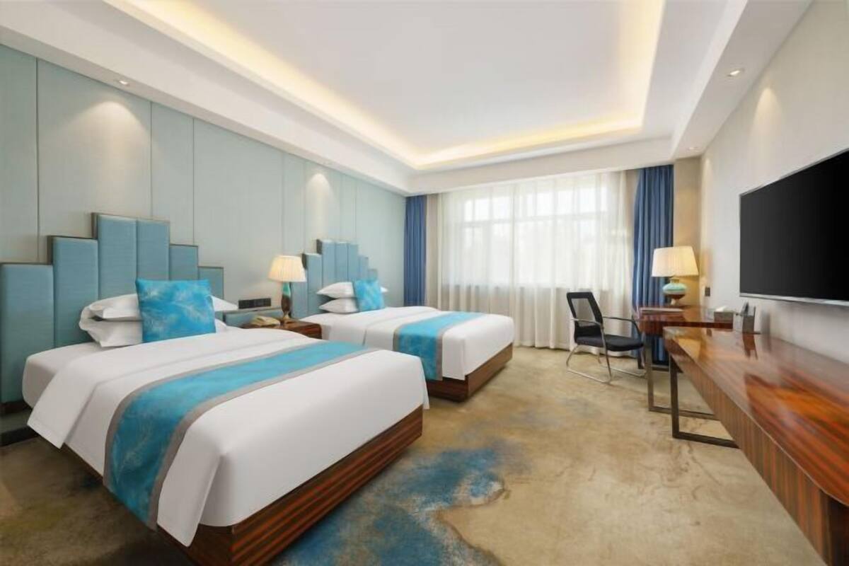 Ein Hotelzimmer mit zwei Betten, einem Schreibtisch, einem Fernseher und einem großen Fenster mit Vorhängen.