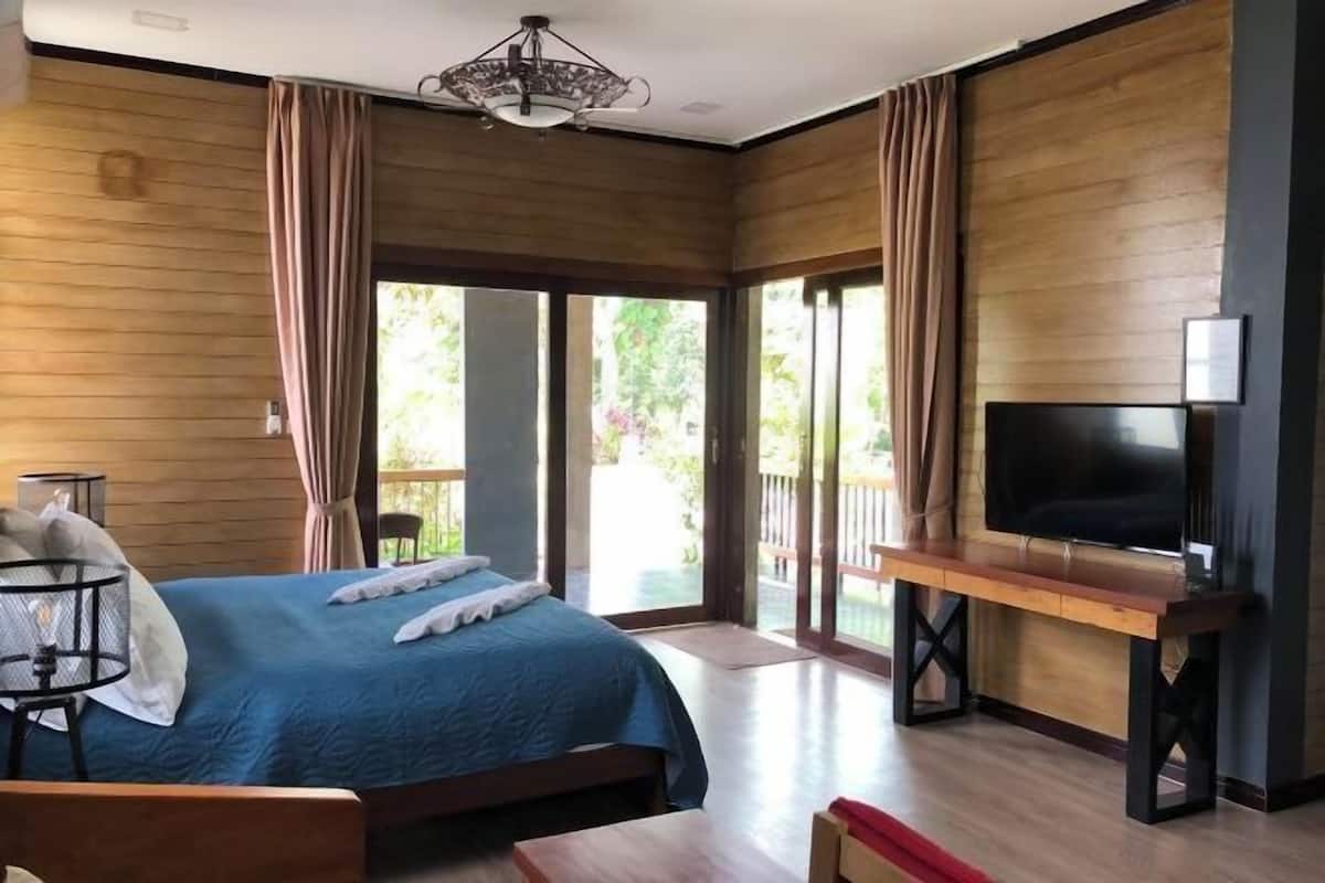 A hotel room with a large bed, a TV on a wooden stand, and a balcony with curtains.