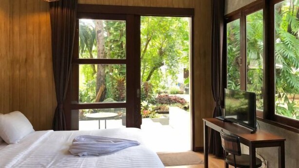 A bedroom with a bed, a TV on a desk, and a view of greenery outside. - Likita Resort (Rayong City)