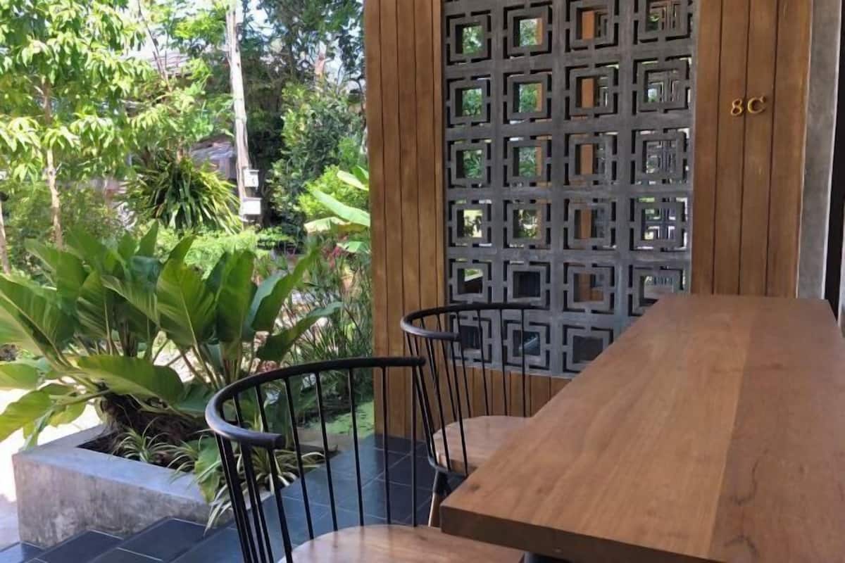 A wooden table and two chairs in a garden area with a decorative metal screen and a number '8C' on the wall.