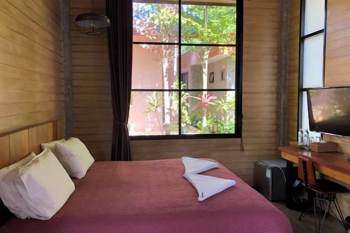 A hotel room with a bed, a desk, a TV, and a window with a view of greenery.