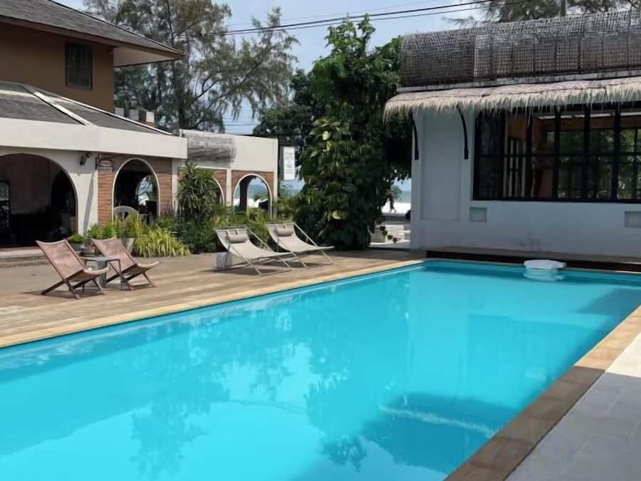 Una piscina con sillones de descanso, una estructura con techo de paja y un edificio con puertas arqueadas.