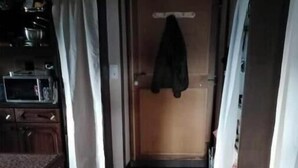 Ein Zimmer mit einem Holzkasten, einer Mikrowelle, einer Tür mit aufgehängter Kleidung und einer roten Matte auf dem Boden.