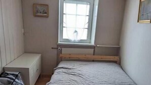 Ein kleines Zimmer mit einem Einzelbett, einem Holzkopfteil und einem Fenster mit Blick auf eine Person draußen.