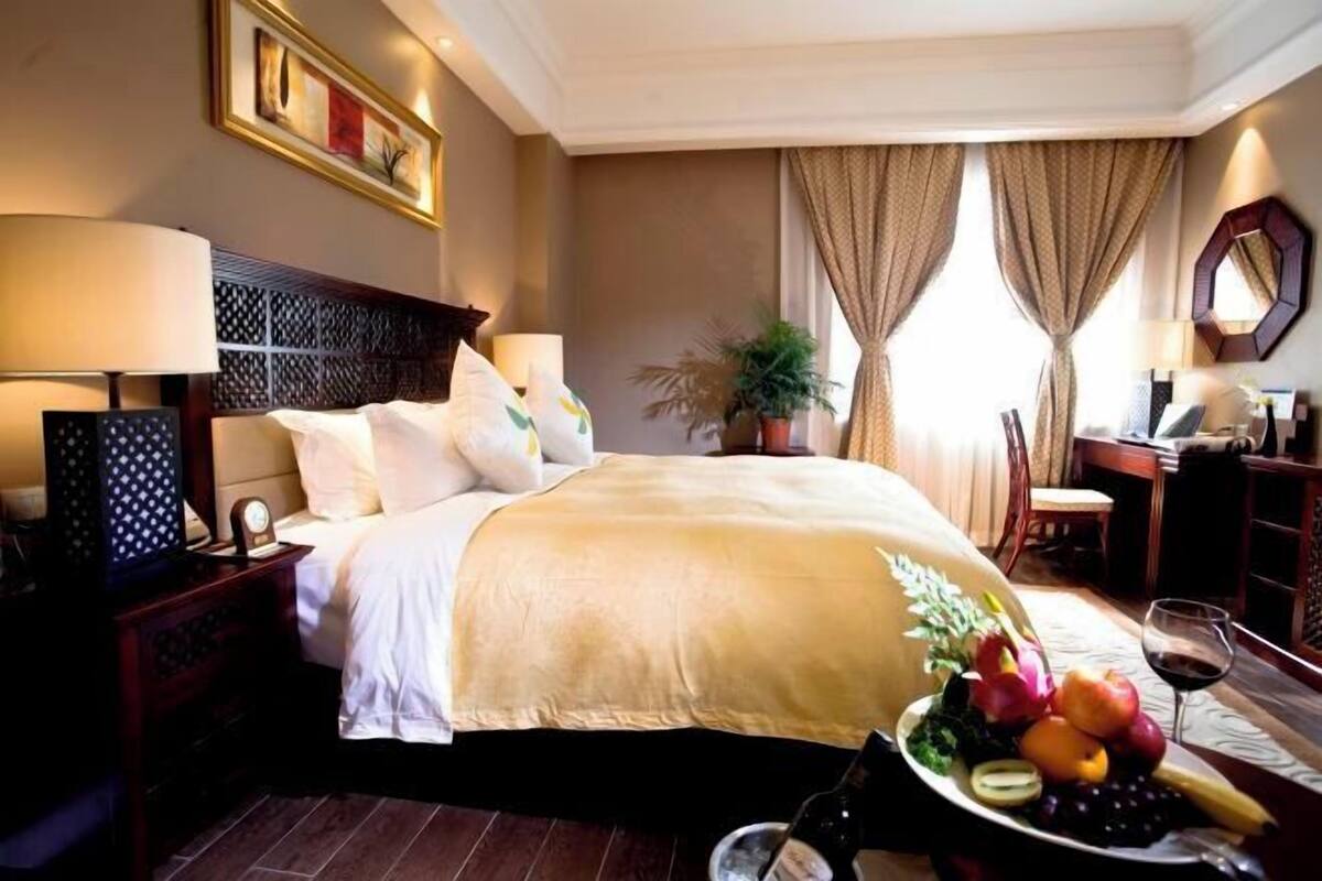 A hotel room with a bed, bedside tables, a desk, a chair, a potted plant, and a fruit basket.